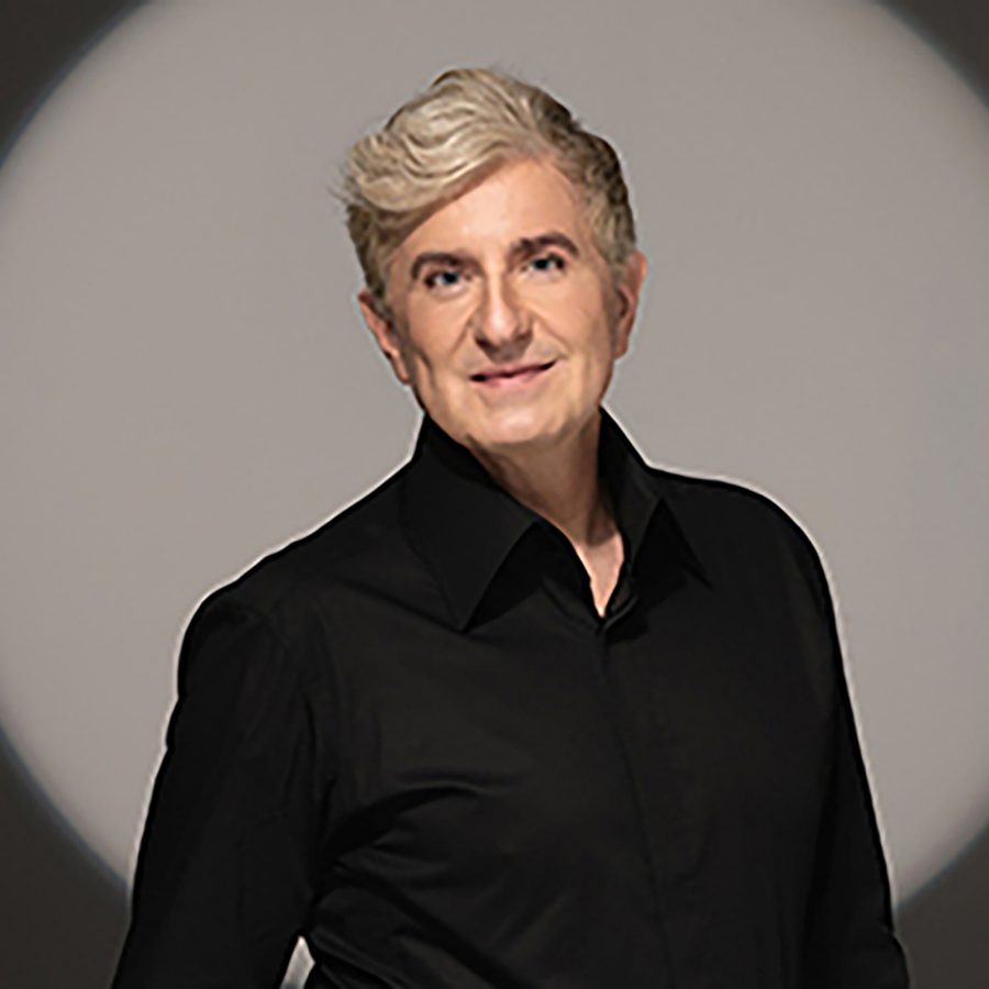 A portrait photo of French pianist Jean-Yves Thibaudet, dressed in a black shirt and standing against a plain background, illuminated by a spotlight.