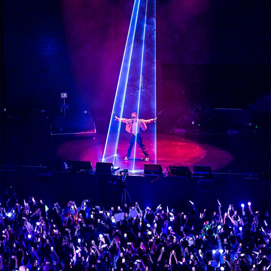 Singer Rain poses on a stage surrounded by blue strobe lights. Below the stage spectators shine phone flashlights.