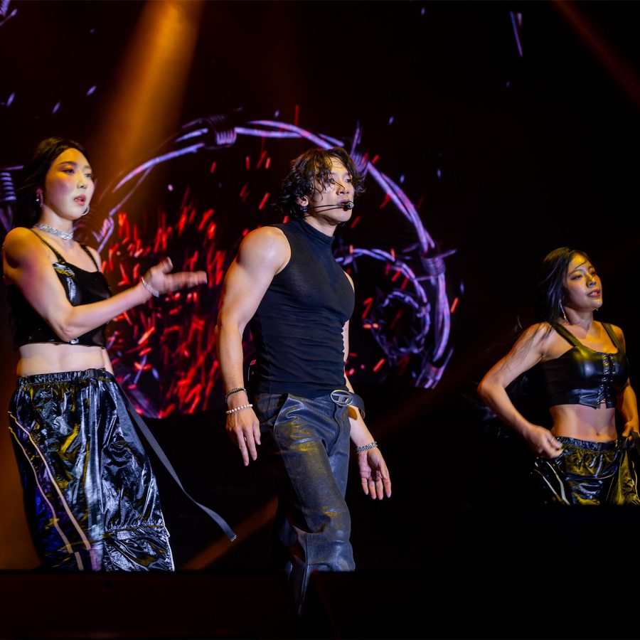 Singer Rain performs on stage wearing a black leather outfit, accompanied by two female dancers dressed in black PVC outfits. 