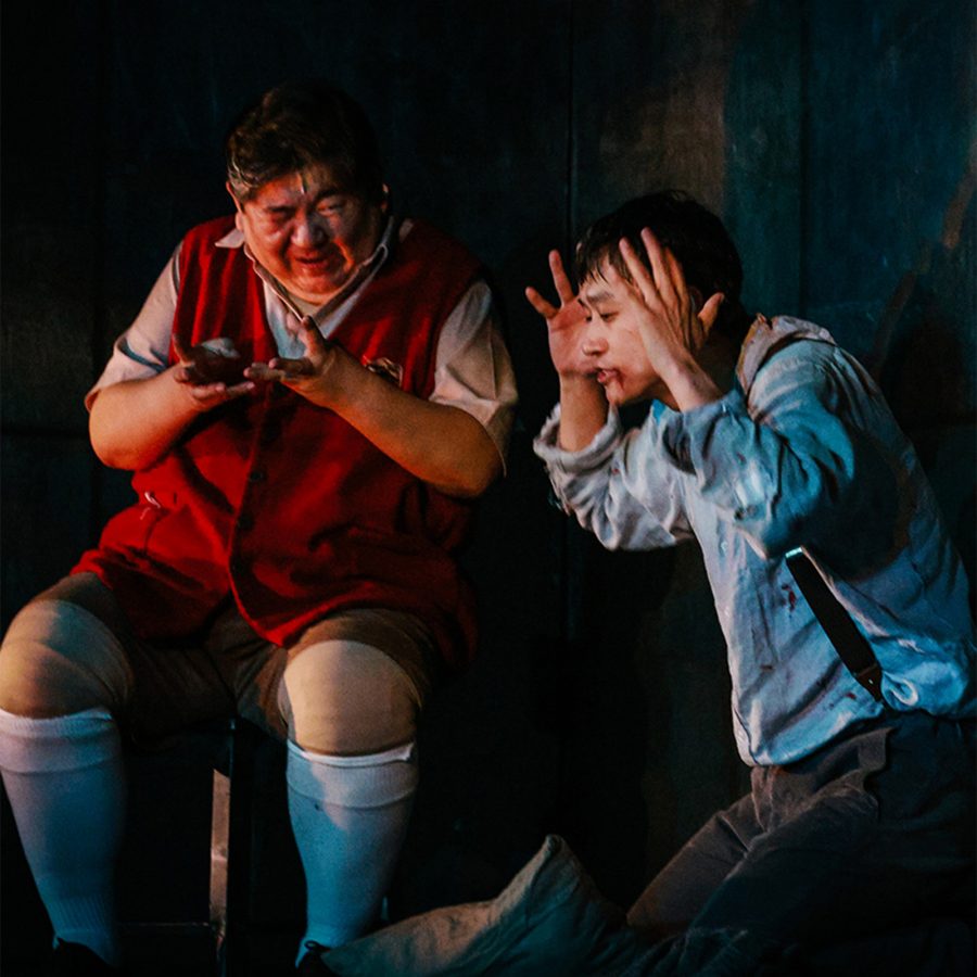 A scene from the stage play The Pillowman shows two men with bloody facial injuries seated against a dark backdrop. 