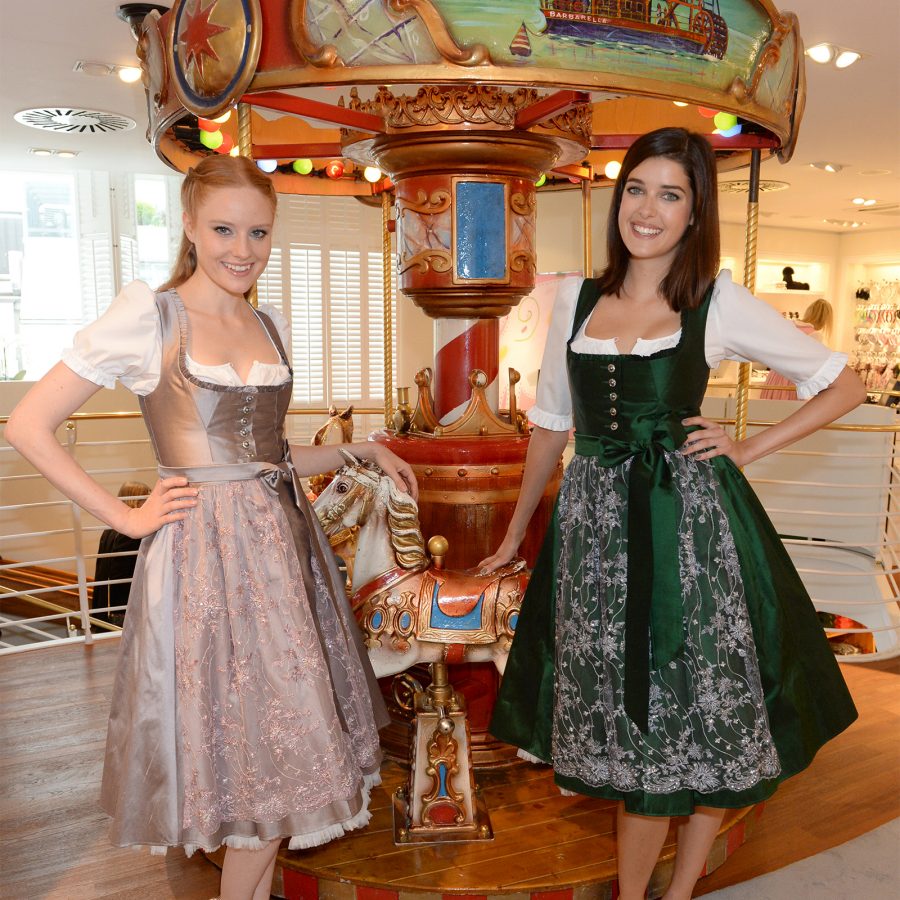 Two women wear traditional dirndls beside a wooden carousel. 