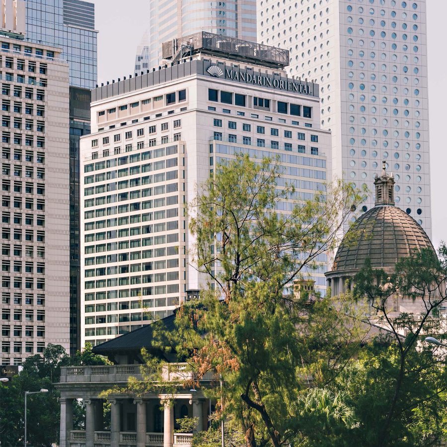 Exterior of Mandarin Oriental Hong Kong building, seen from behind heritage buildings and trees, in daytime. 