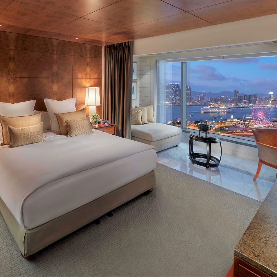 A hotel room with a king-size bed against a wood panelled wall, with glowing lamps either side. Curtains separate the bed from a sitting area and floor-to-ceiling window that looks onto Victoria Harbour. 