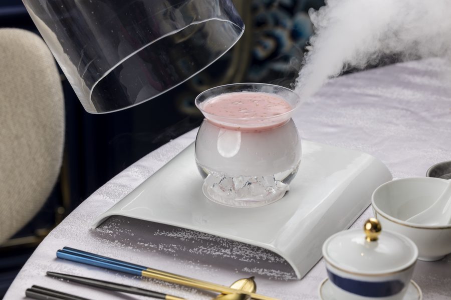 A stream of water vapour rises from a glass bowl of pink and cream dessert, displayed on an elegantly laid table. 