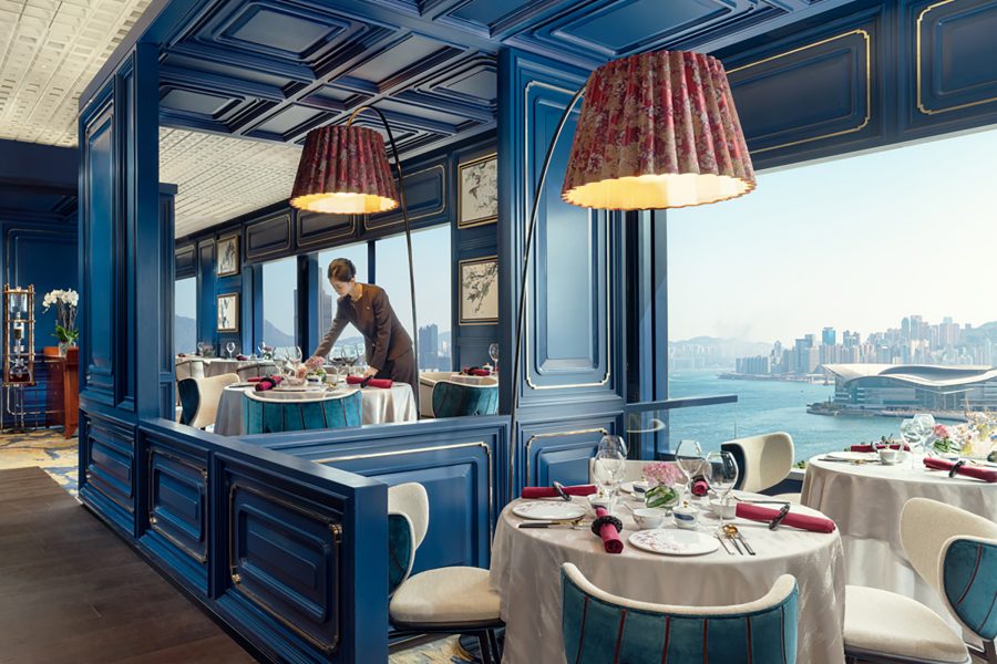 The interior of Mah Wah, featuring blue walls, elegantly laid round tables, teal and white chairs and statement lamps. A staff member arranges a table setting. 