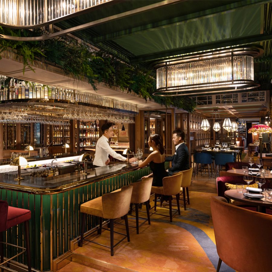  The interior of The Aubrey, featuring Art Deco lighting and lounge seating. A couple sits at a green and gold bar, being served by a bartender in a white shirt and waistcoat. 