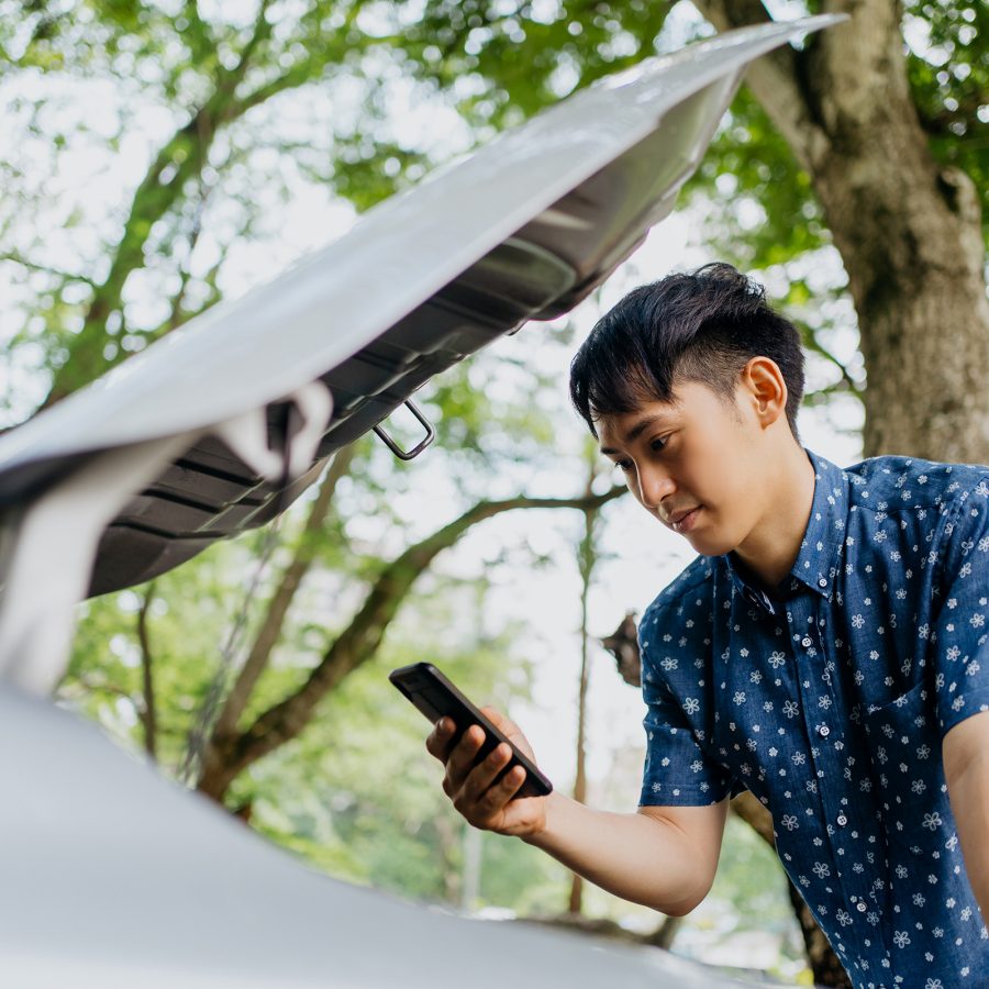 A man checks his phone while standing in front of his car. The hood of the car is open.