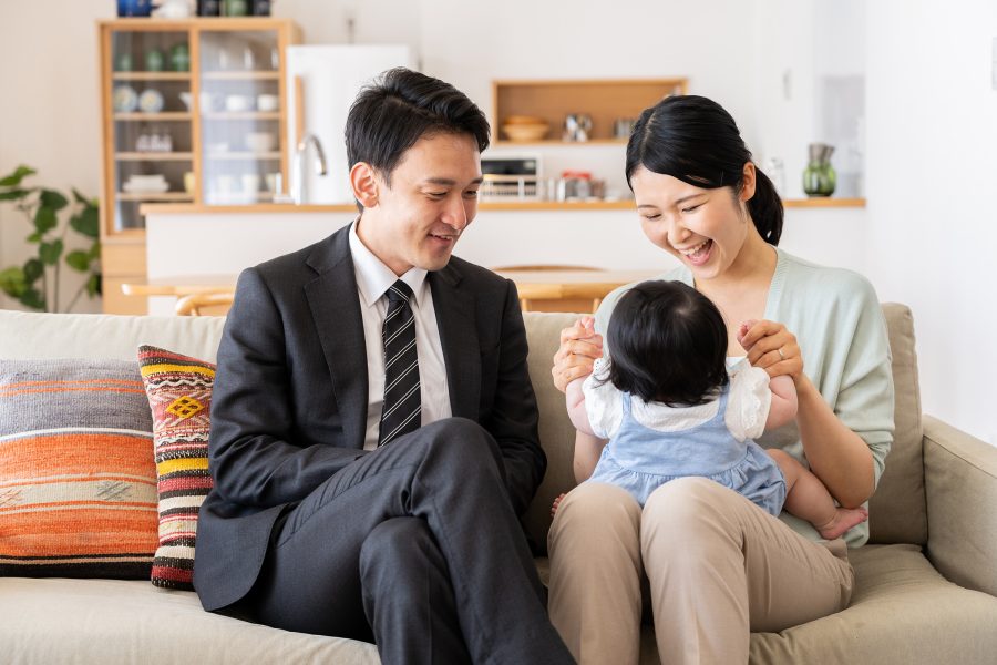 A man and a woman with a baby sit on a beige sofa in a living room.