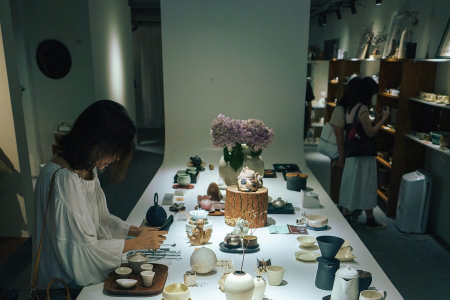A woman sitting at a table taking a photo of ceramic works at OCT Loft creative park while two women browse the shelf in the background.