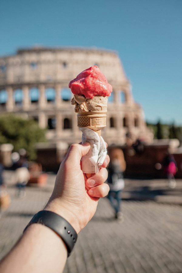 A tourist holds up an ice cream cone with a pink scoop of ice cream in front of the Colosseum. 