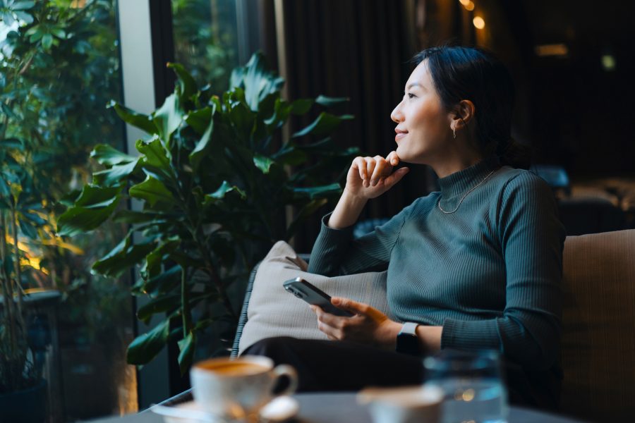 A woman sitting on a couch with her phone in one hand, looking out a window.