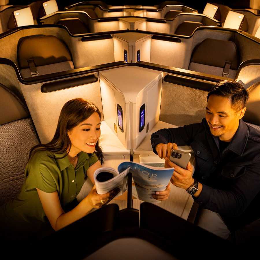 A man and a woman sitting on a plane. The woman is showing the man her in-flight magazine while the man is looking at his phone. 