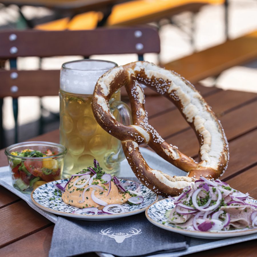A closep of a large pretzel bread leaning on a tall beer galss over small dishes at Hirschgarten in Munich, Germany