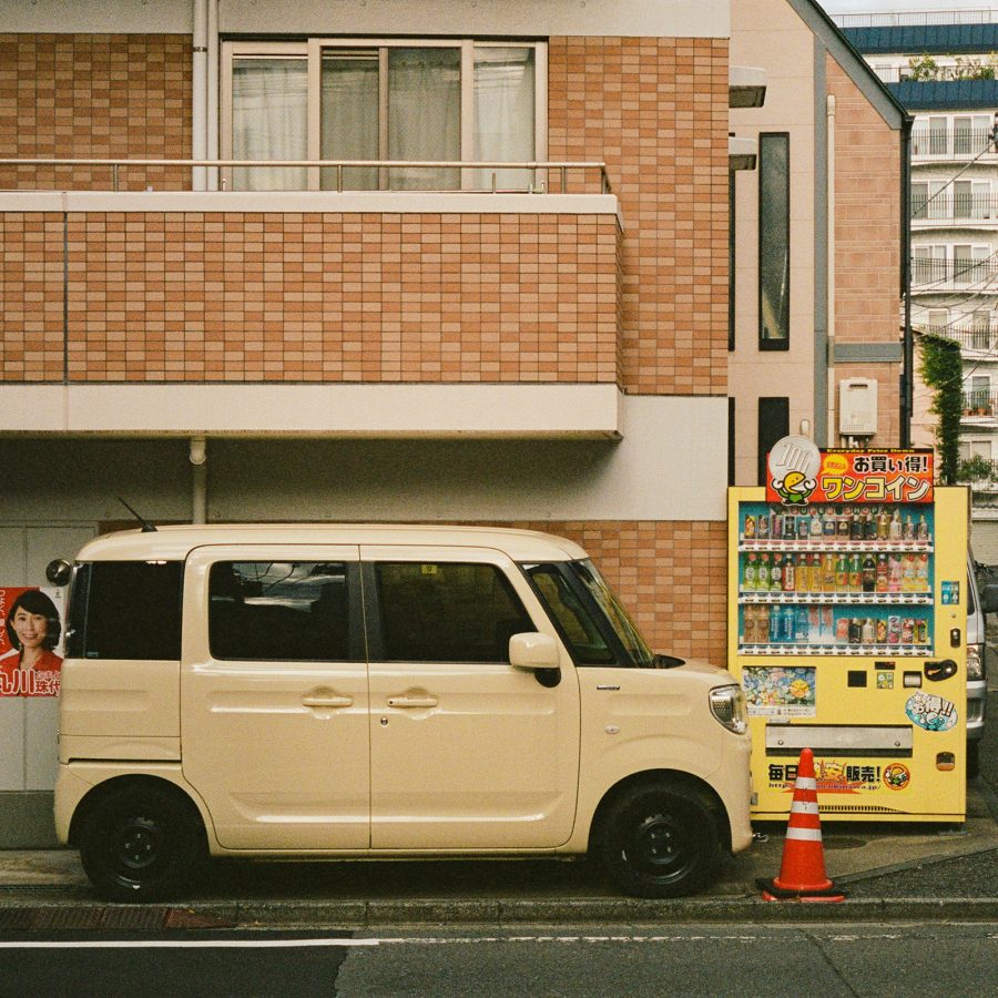 Boxy Japanese car parked beside a yellow vending machine.  