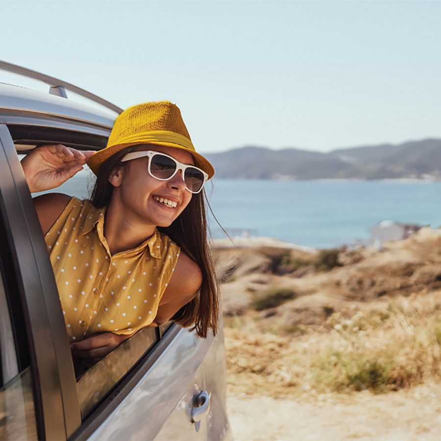 A woman in a yellow shirt, hat and sunglasses smiles out of a car window on a sunny day. Hills and a body of water are in the background.