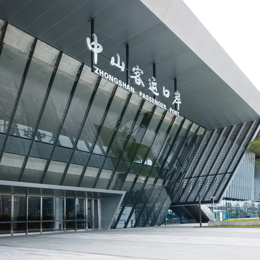  The exterior of the Zhongshan Passenger Port is a tall grey building with slanted glass walls and a giant sign above. 