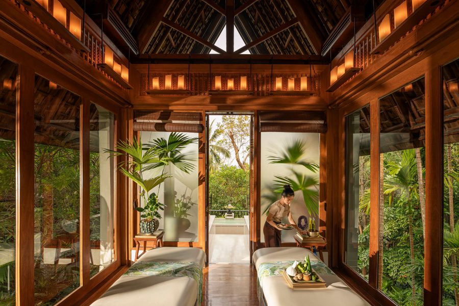 A staff member inside a spa treatment room at Four Seasons Koh Samui with wooden interiors, two beds and the forest landscape through the windows. 