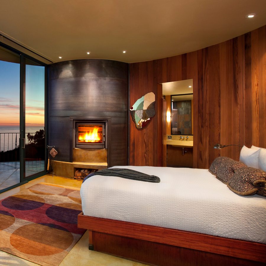 A curved room at Post Ranch Inn with a fireplace, bed, table and a sunset view through the floor-to-ceiling window.
