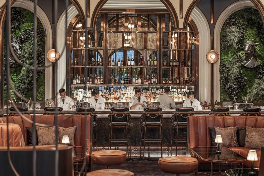 An interior view of the luxurious BKK Social Club in Bangkok, featuring elegant low-lit lamps, sleek leather booths, tropical greenery on the walls and five mixologist working behind the bar