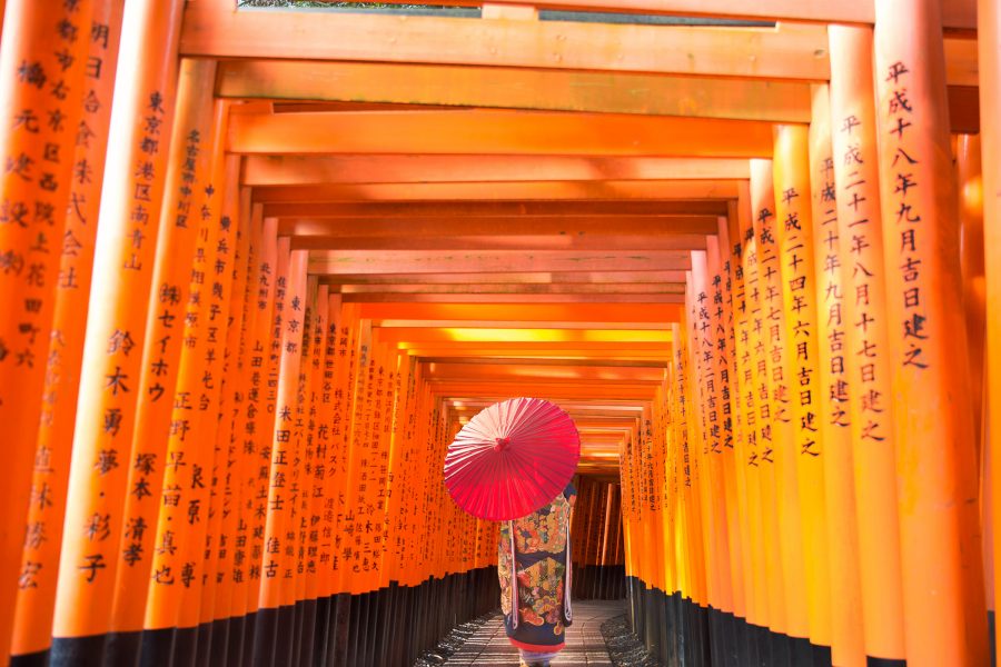 A person dressed in traditional Japanese clothing and holding an umbrella, standing in the middle of a torii path at Fushimi Inari Shrine, Kyoto.