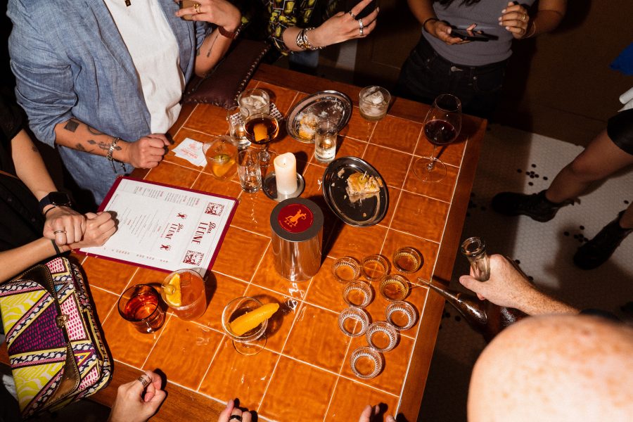 People gather around a wooden table filled with drinks and small bites at Bar Leone.