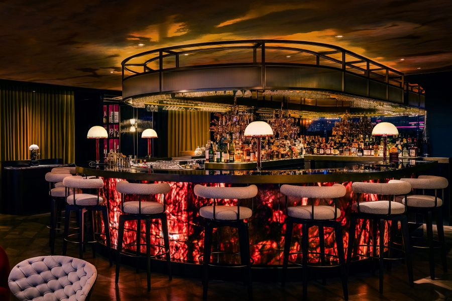 The bar at Qura glows red, surrounded by white-cushioned bar stools and Art Deco-inspired décor.