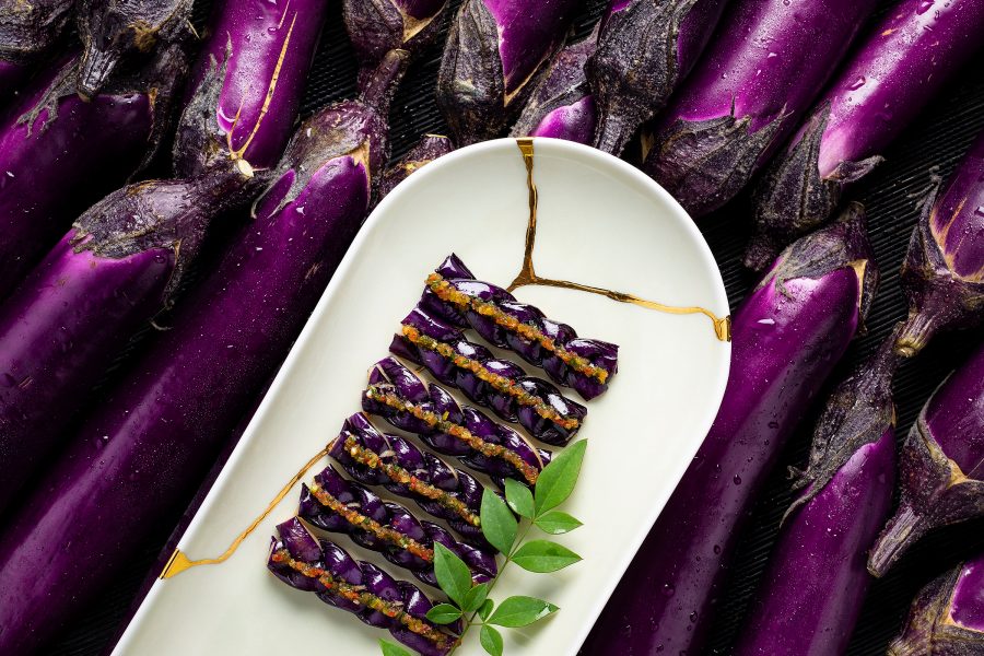 A white dish from Wing, filled with slices of seasoned eggplant, rests on top of two rows of the purple vegetable.