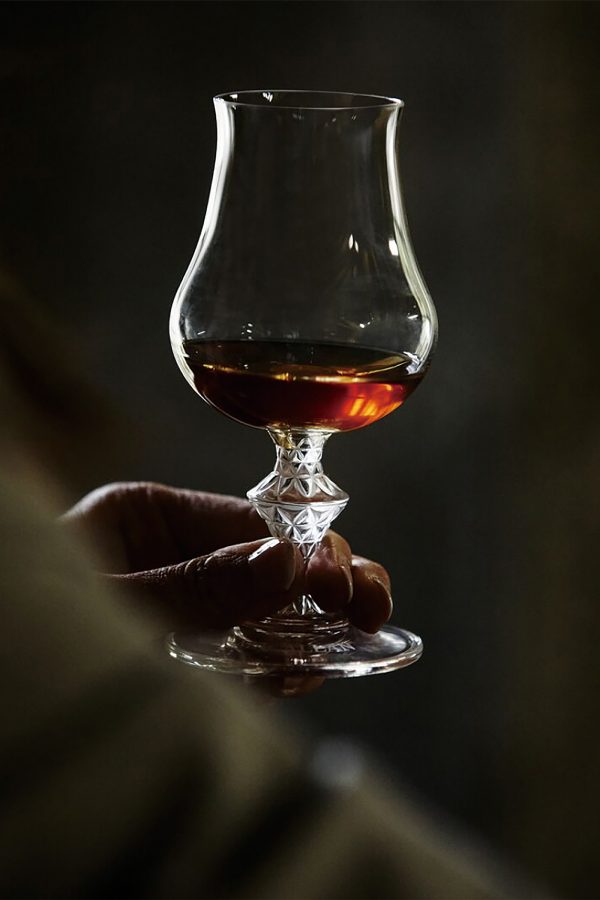 Person holds up a dram of whisky in a crystal glass. 