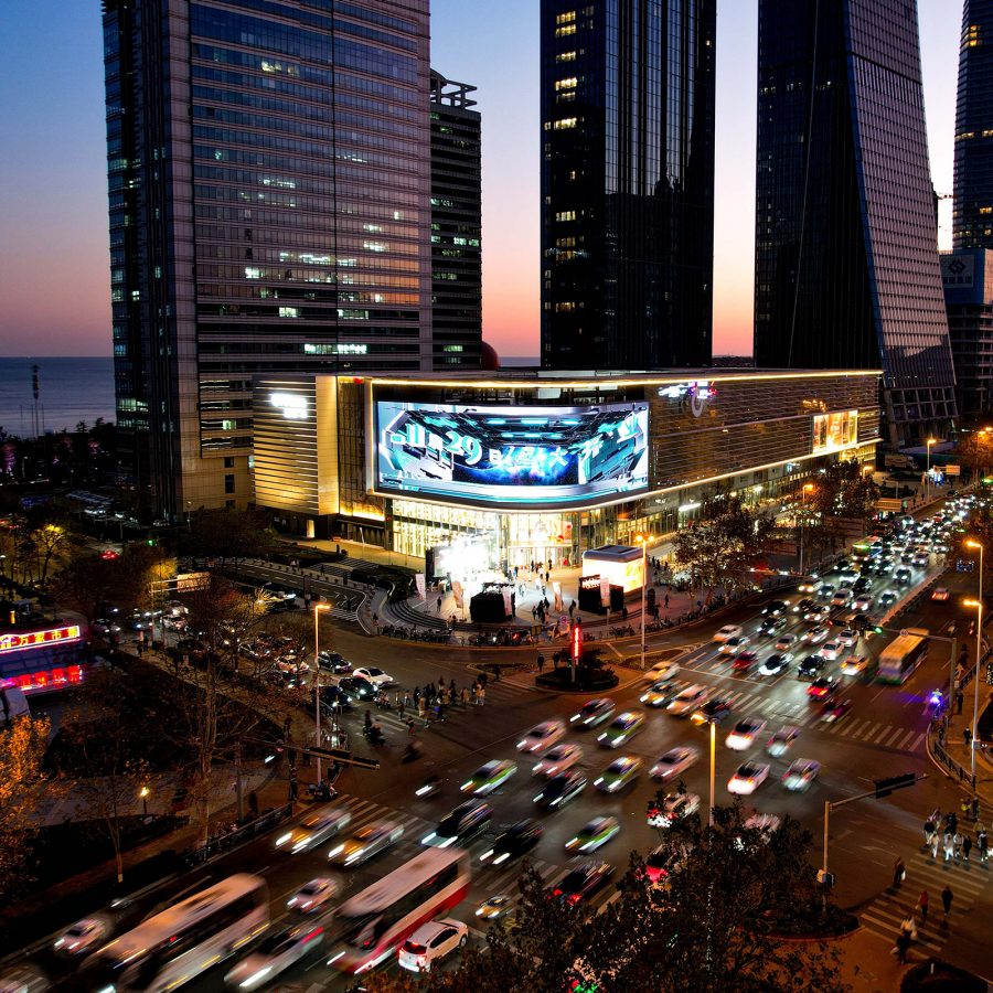 Exterior view of GT Plaza Qingdao shopping mall by night, with water in the background and cars in the foreground.