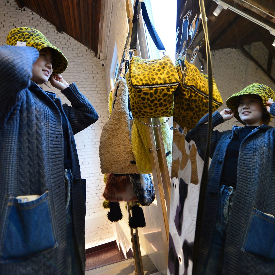 A young woman stands in front of a shop mirror and tries on a yellow-and-black polka dot hat on Silverfish Lane, Qingdao. 