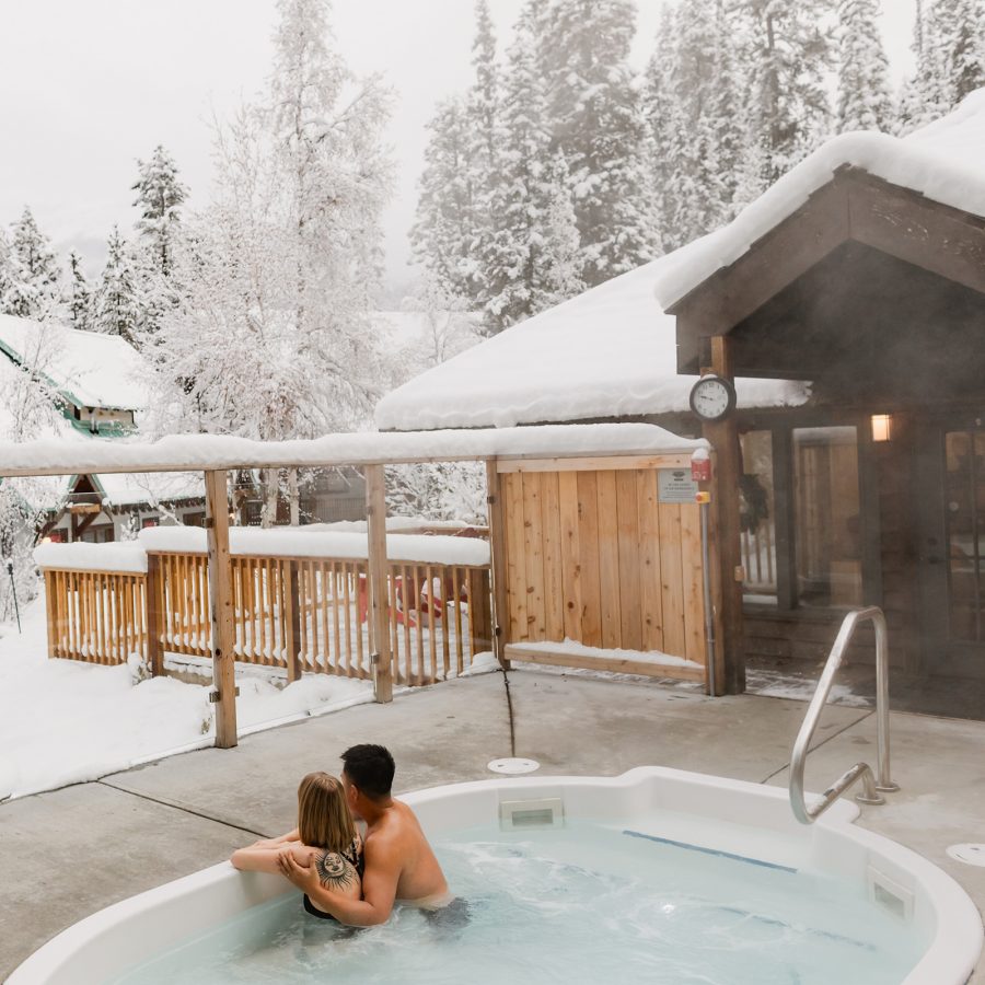 Exterior view of a couple enjoying a jacuzzi together as they look at snow-covered pine trees.
