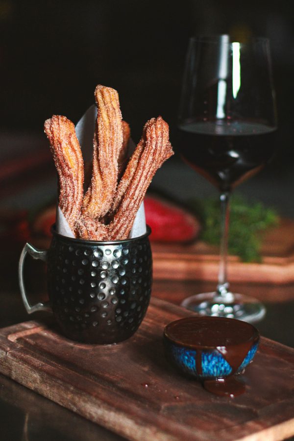 A mug holds a handful of churros, next to a dip and a glass of wine at Picanhas’. 