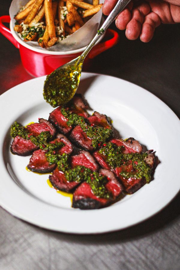 A hand spoons dollops of chimichurri over red slices of wagyu at Picanhas’, next to a dish of fries. 