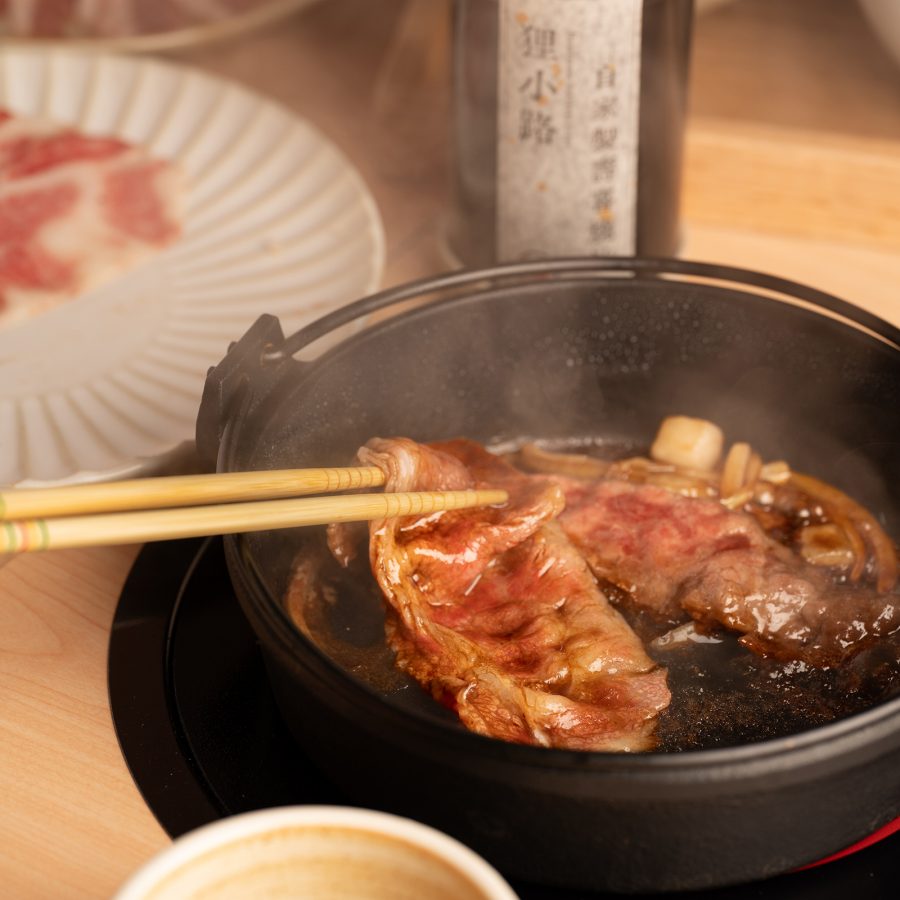 An offscreen diner enjoying an order of shabu-shabu cooks slices of beef on a sizzling pot. 