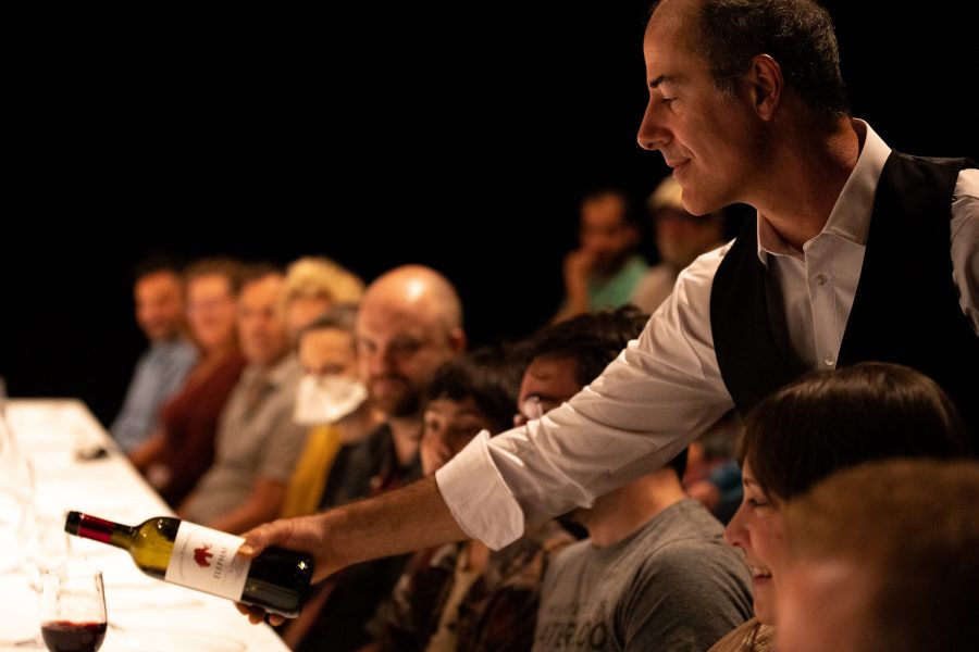 Geoff Sobelle pours wine from a bottle for a long table of people. 