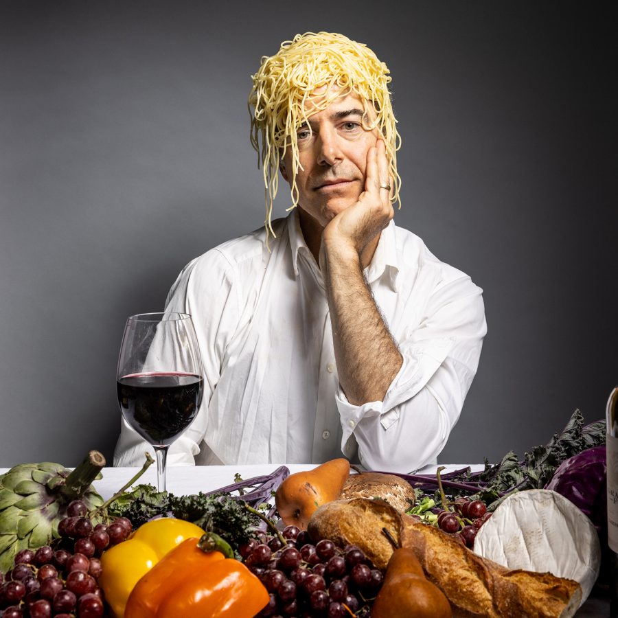 Geoff Sobelle sits musingly behind a table filled with food, with a bunch of spaghetti on his head.