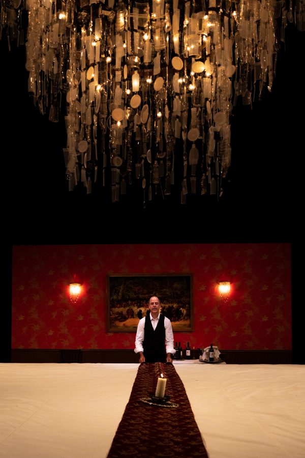 Geoff Sobelle stands behind a large table in front of a deep red wall as a chandelier hangs down from the ceiling. 