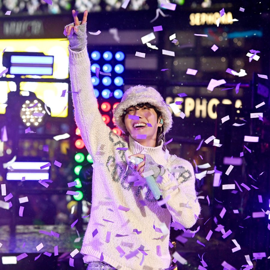 J-Hope raises an arm showing the peace sign onstage as lights and signs glow behind him. 
