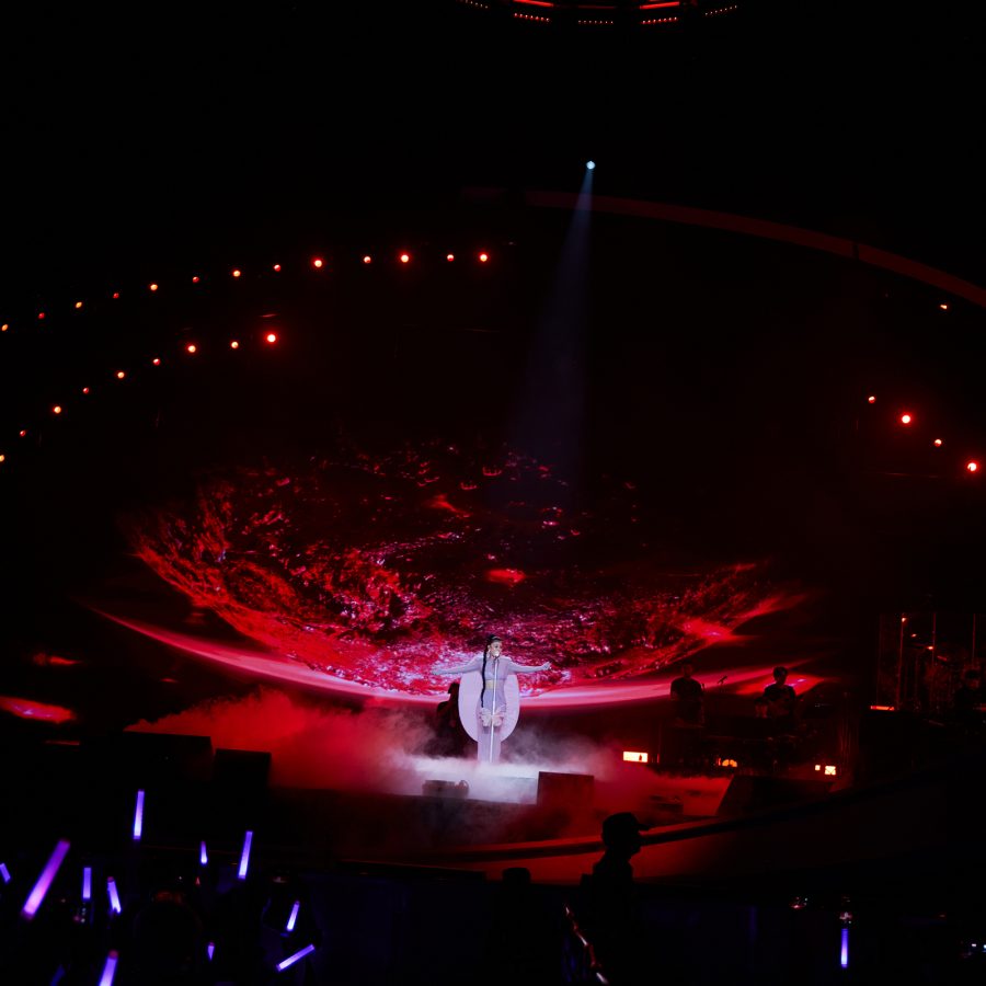 Tia Ray stands on the stage as the backdrop behind her glows red and light sticks are raised toward the stage.