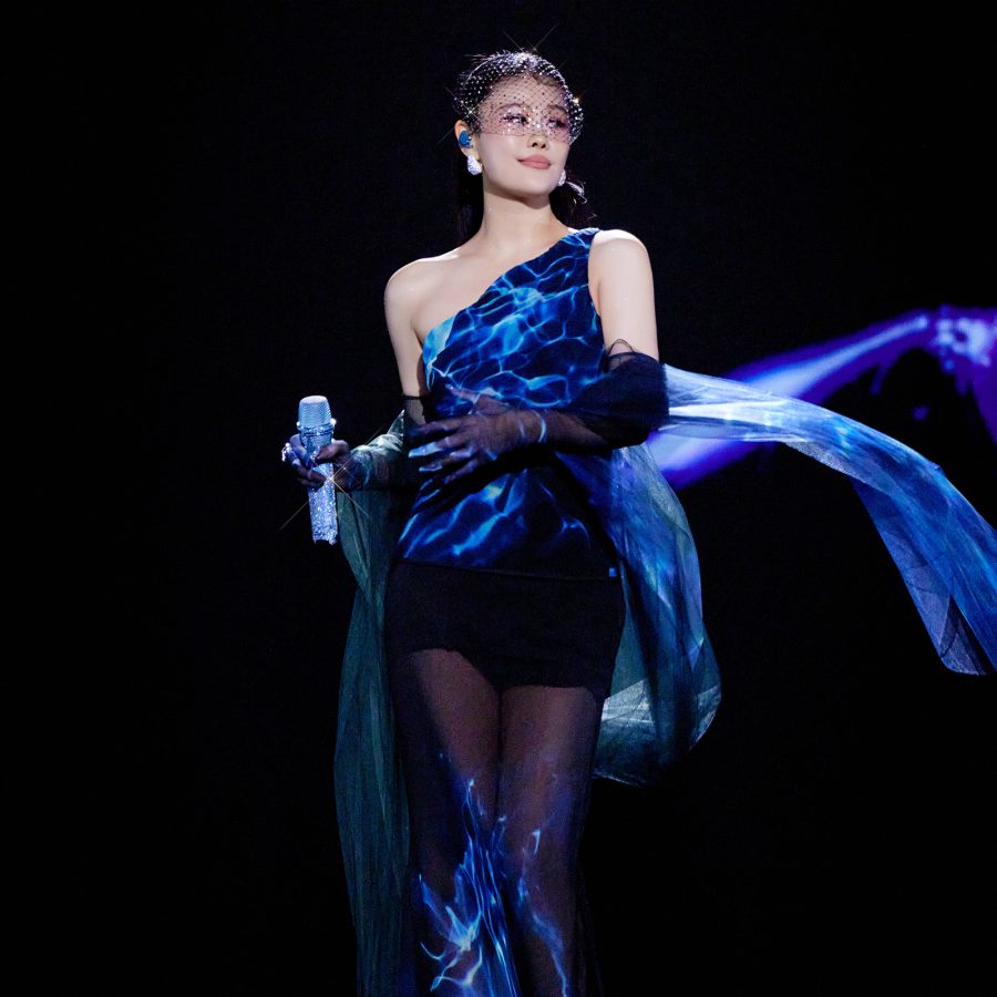 Tia Ray stands on stage with a microphone in hand in a flowy blue dress. 