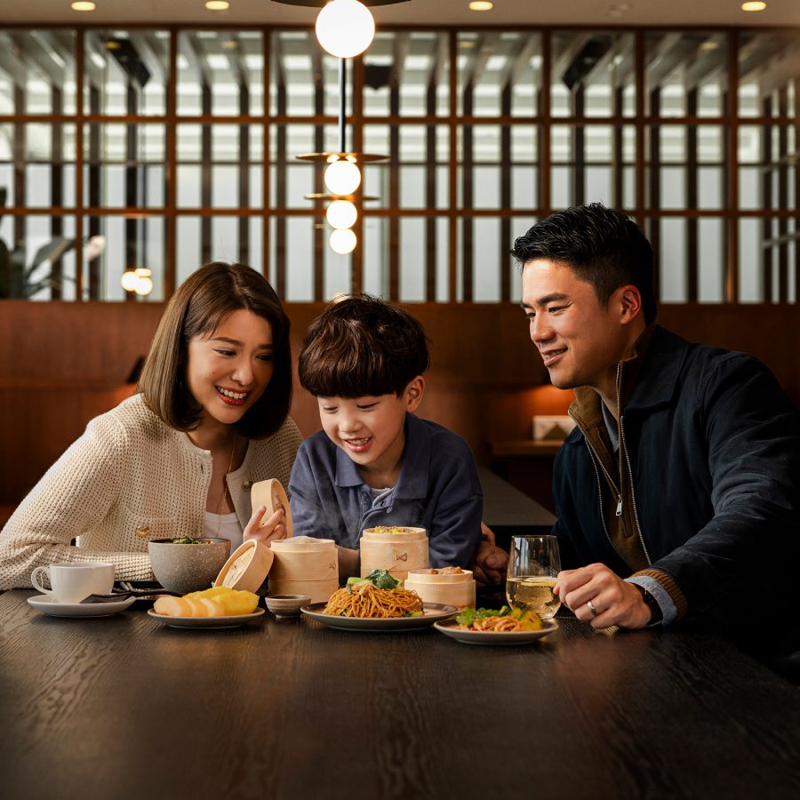 A smiling family of three sit at a table in The Bridge with various dishes in front of them, including dim sum and noodles. 