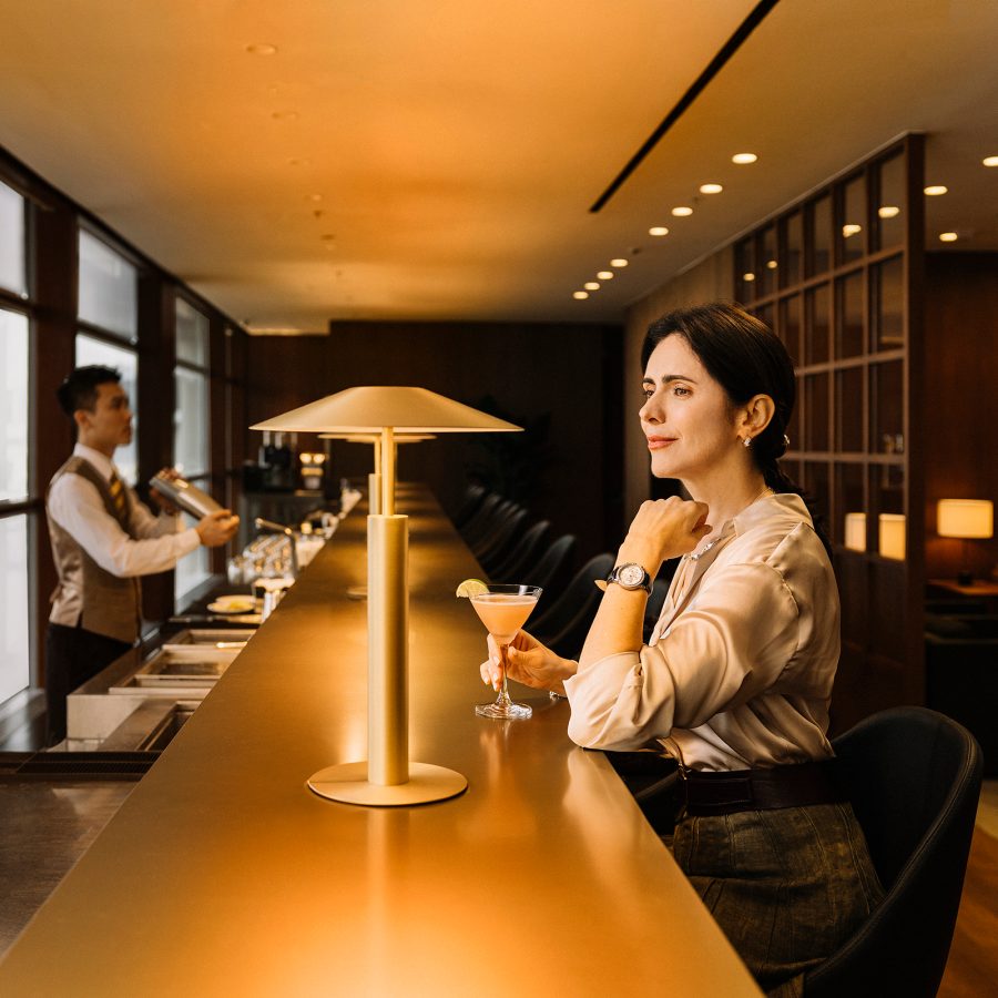 A customer sits at The Long Bar with a drink in hand as she looks out the window, as the bartender prepares another drink behind the counter. 