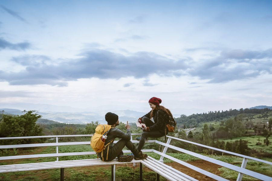 Man wearing a travel backpack talks to a woman with a journal in her hands as they sit in front of a grassy landscape.