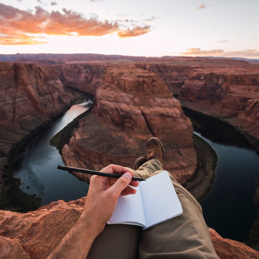 Person writes in an empty journal with a view of a natural rock formation and lake below.