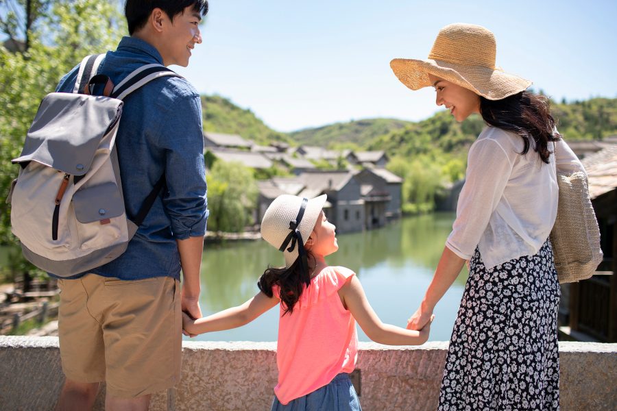 Exterior of countryside with a man, a woman and a young girl standing on a bridge overlooking a body of water, with old buildings in the background. 
