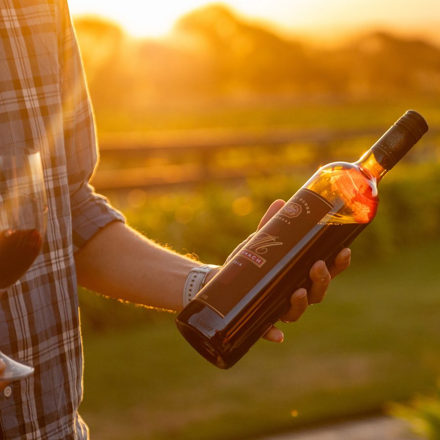 A closeup of hands holding a bottle of Grant Burge and a glass of red wine with a view of the sun setting over the vineyard in the background.