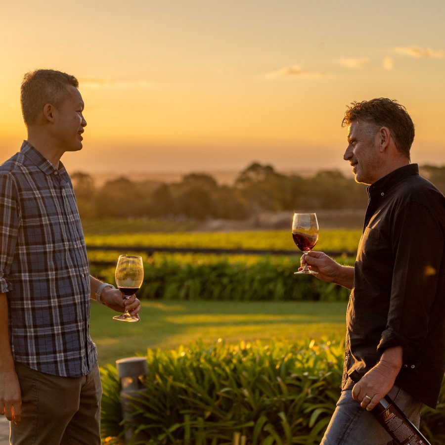 Cathay Pacific’s Ronald Khoo and Vinarchy’s Craig Stansborough chatting while holding a glass of red wine with a view of the vineyard and sun setting in the background.