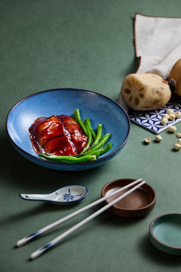 A dish with braised duck in red fermented rice sauce served with greens. Lotus root and seeds, chopsticks and a ceramic spoon are placed around the dish.