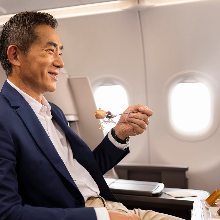 A man sitting in an aircraft cabin smiles as he holds up a spring roll with his fork. 