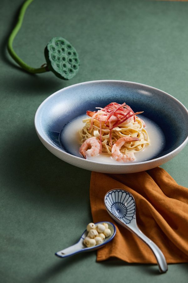 A dish with braised prawns with dried shredded tofu and Chinese ham. A lotus seed pod and ceramic spoons are placed around the dish. 